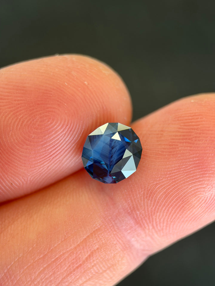 Opalescent royal blue sapphire with natural growth patterns held in fingertips under open shade showing the scale
