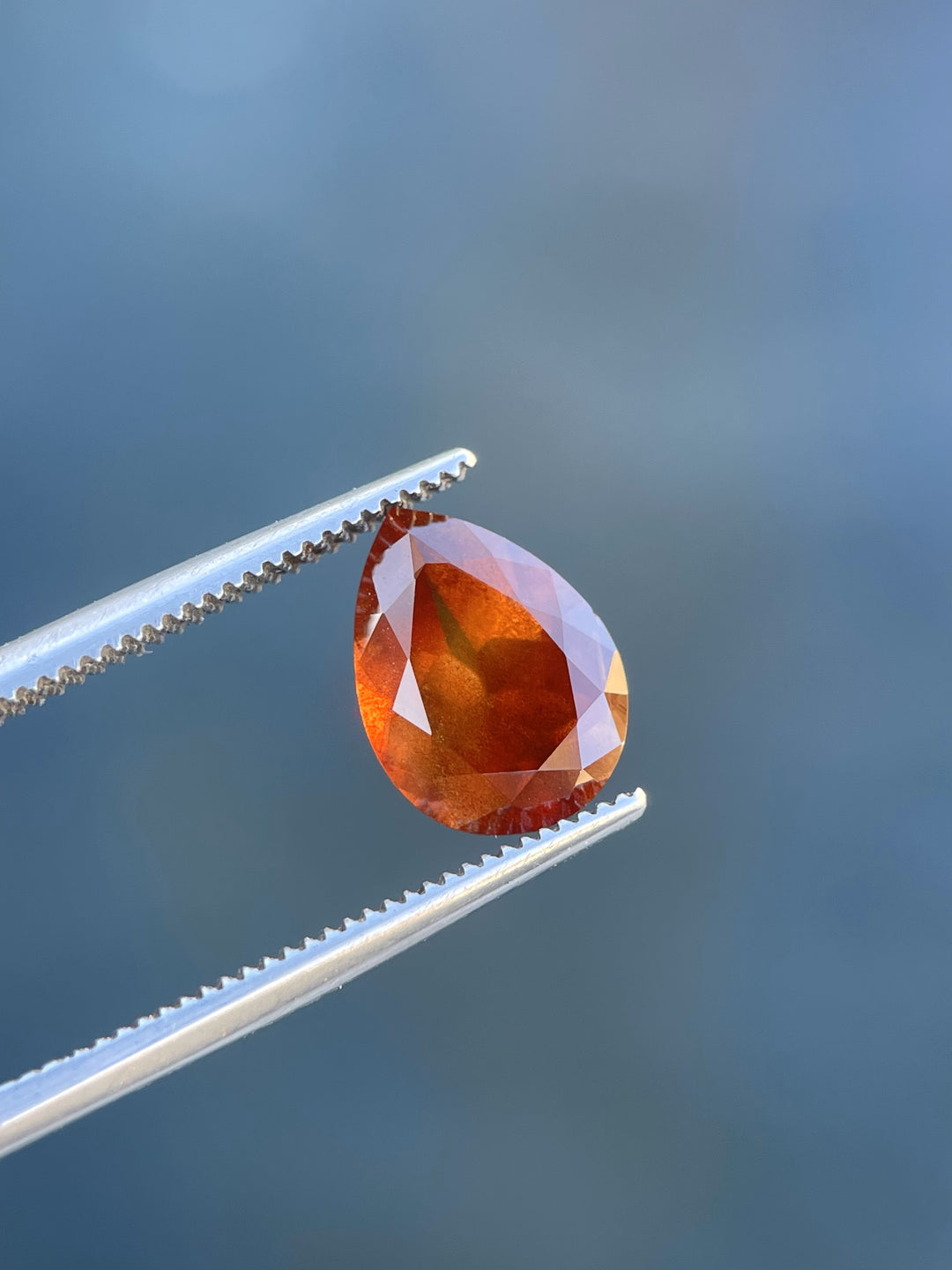 Vibrant orange sapphire gemstone held with tweezers, showcasing its 2.90 carat weight and modified pear cut.