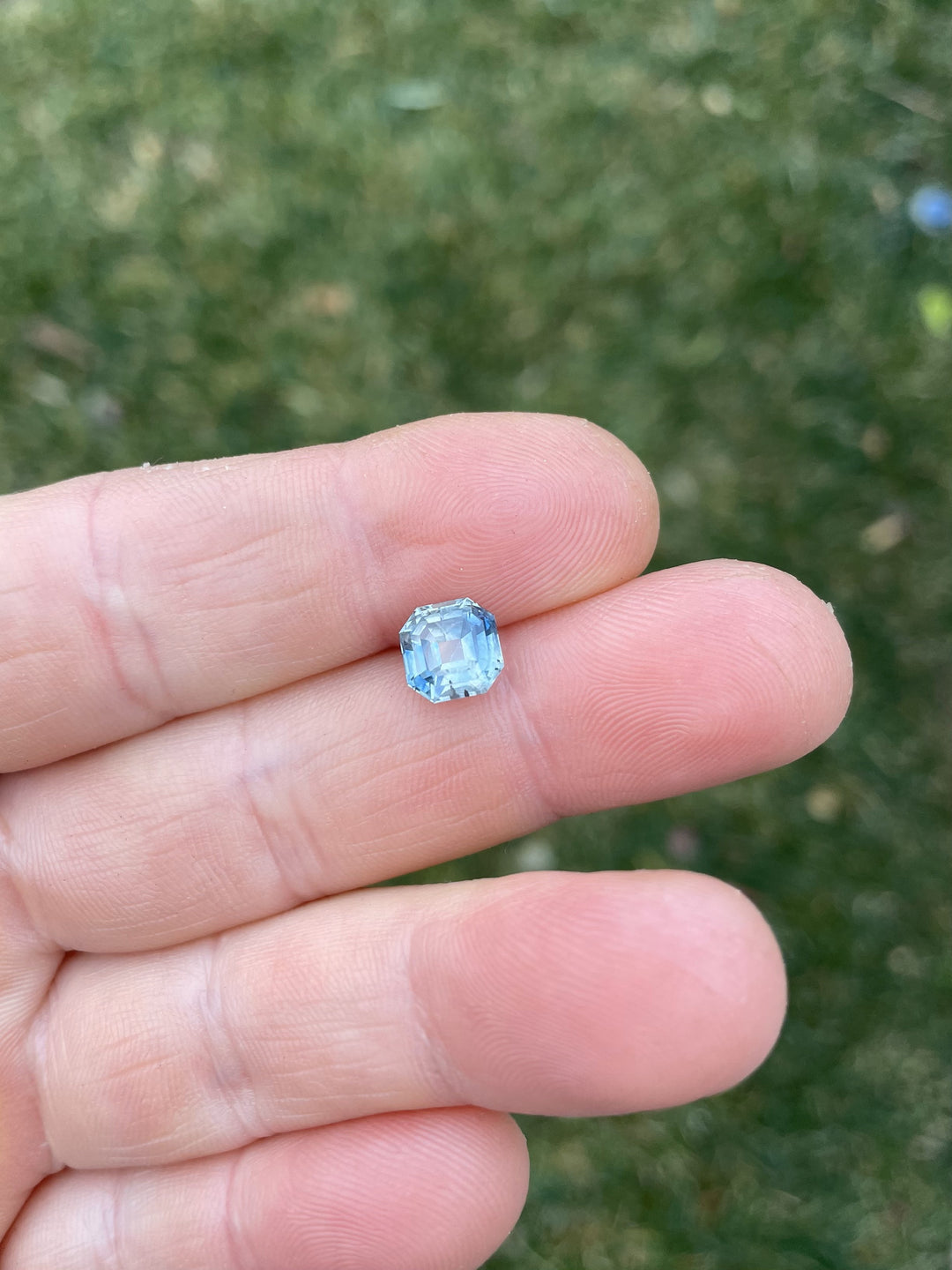2.45-carat parti blue-green sapphire in an antique emerald cut, placed delicately on fingers against a green grassy background, showcasing its proportions and sparkle.