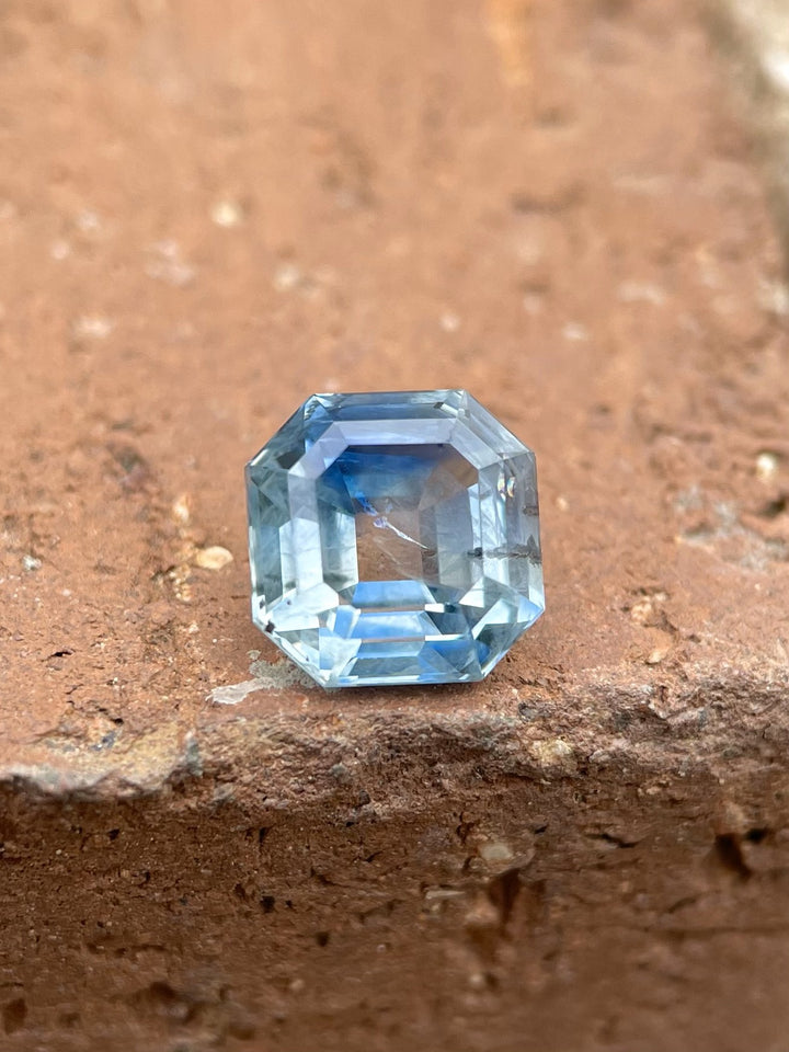 2.45-carat parti blue-green sapphire in an antique emerald cut, resting on a rustic, earthy brown surface, showcasing its exquisite step cut and vivid colors.