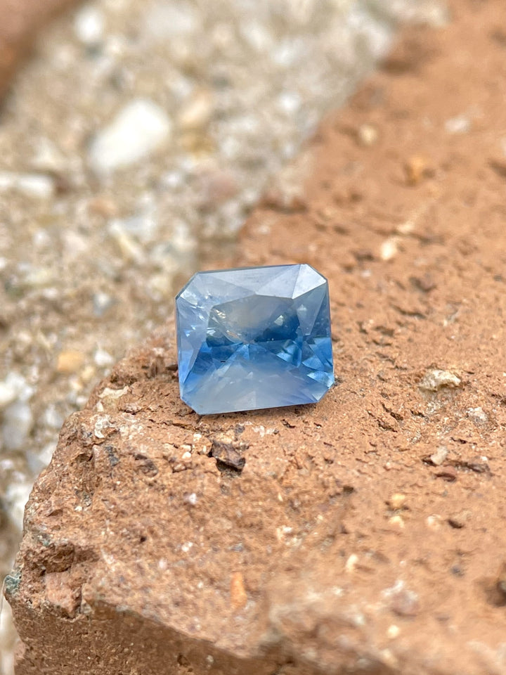 Close-up of a Parti Blue and White Sapphire weighing 1.03 carats, with Mixed Radiant Cut facets reflecting blue and white tones, resting on a textured earthy surface.