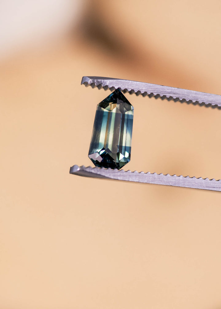 Loose yellow blue green parti sapphire from Madagascar held in tweezers displaying the custom modified step cut shield shape and distinct natural color separation.