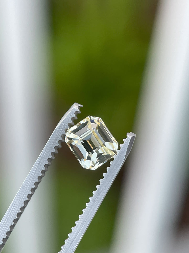 A close-up of a pastel canary yellow sapphire, weighing 1.43 carats, held securely in tweezers. The octagonal step cut facets shimmer beautifully against a blurred green and white background, highlighting its precision and natural elegance.