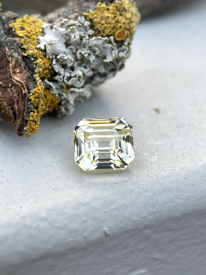 A pastel canary yellow sapphire, weighing 1.43 carats, resting on a lichen-covered surface. The gemstone’s octagonal step cut facets reflect light, showcasing its soft, pastel canary yellow tone and exquisite clarity.