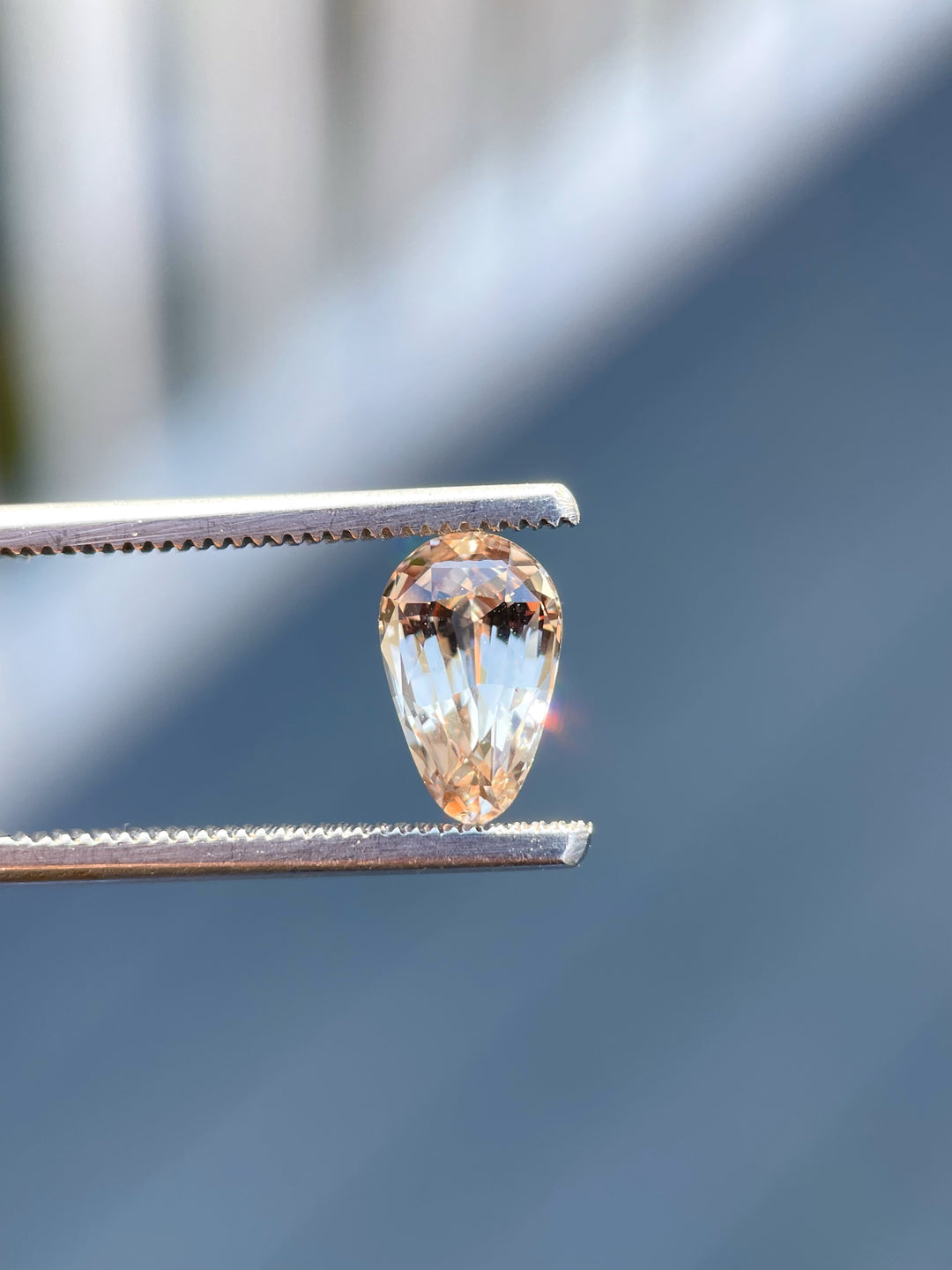 A close-up shot of a 2.45-carat custom brilliant teardrop sapphire held delicately by precision tweezers. The gemstone showcases a pastel champagne and peach color, reflecting subtle light under natural daylight. The gem's dimensions are 10.12mm in length, 6.40mm in width, and 4.70mm in height. Originating from Sri Lanka, this sapphire is untreated and valued at $2,082.50.