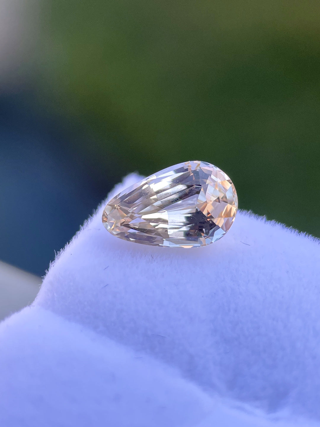A 2.45-carat custom brilliant teardrop sapphire, with soft pastel hues of champagne and peach, rests on a white velvet display. The sapphire's faceted surface captures a delicate shimmer under sunlight. The gemstone measures 10.12mm in length, 6.40mm in width, and 4.70mm in height, ethically sourced from Sri Lanka and untreated.