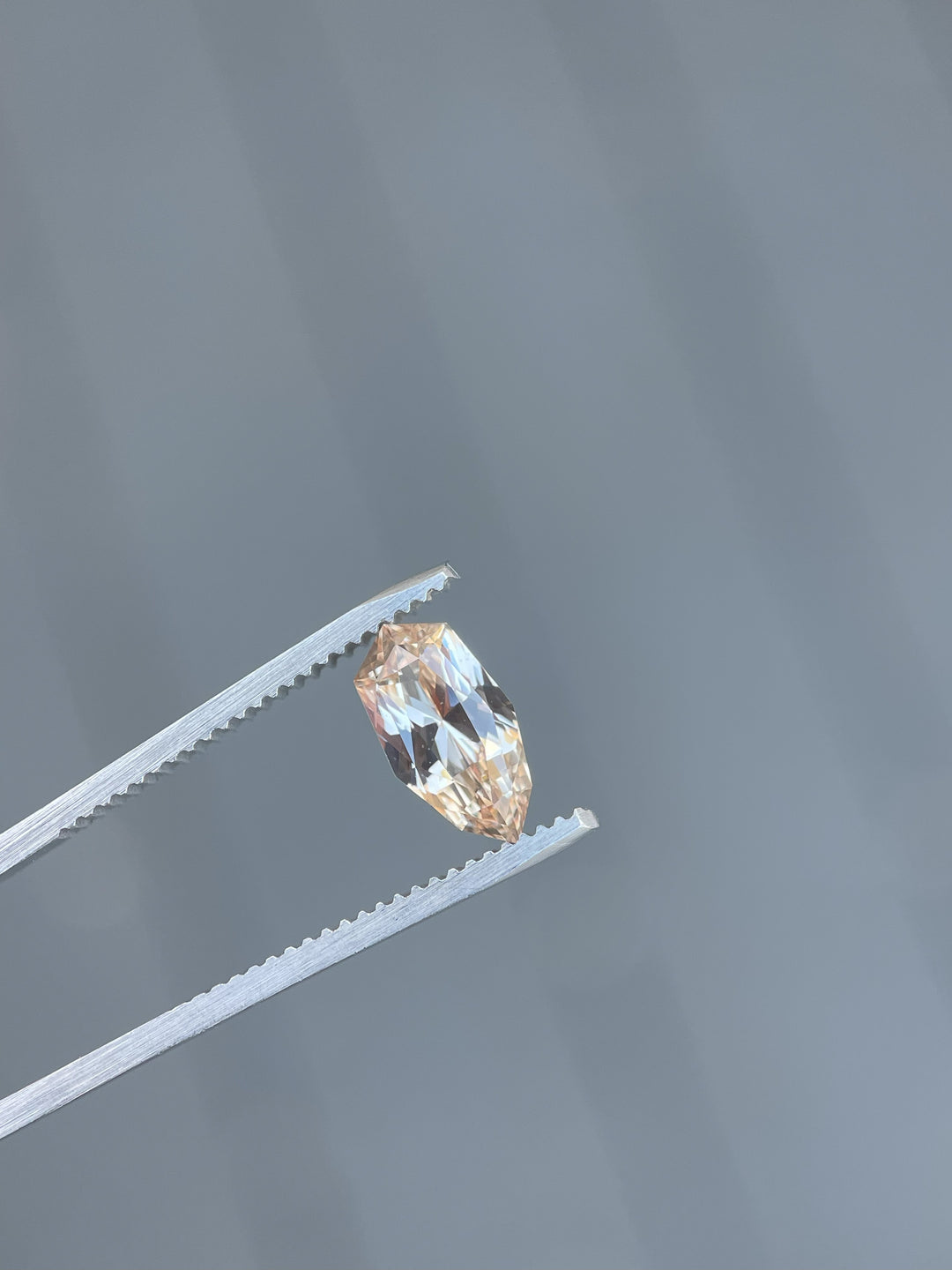 A 2.07-carat peach and champagne sapphire held by precision tweezers. The gemstone, cut in a custom geometric shield brilliant cut, shows its light peach and champagne hues under natural light. It measures 11.02mm by 5.87mm by 3.94mm and originates from Sri Lanka, untreated.