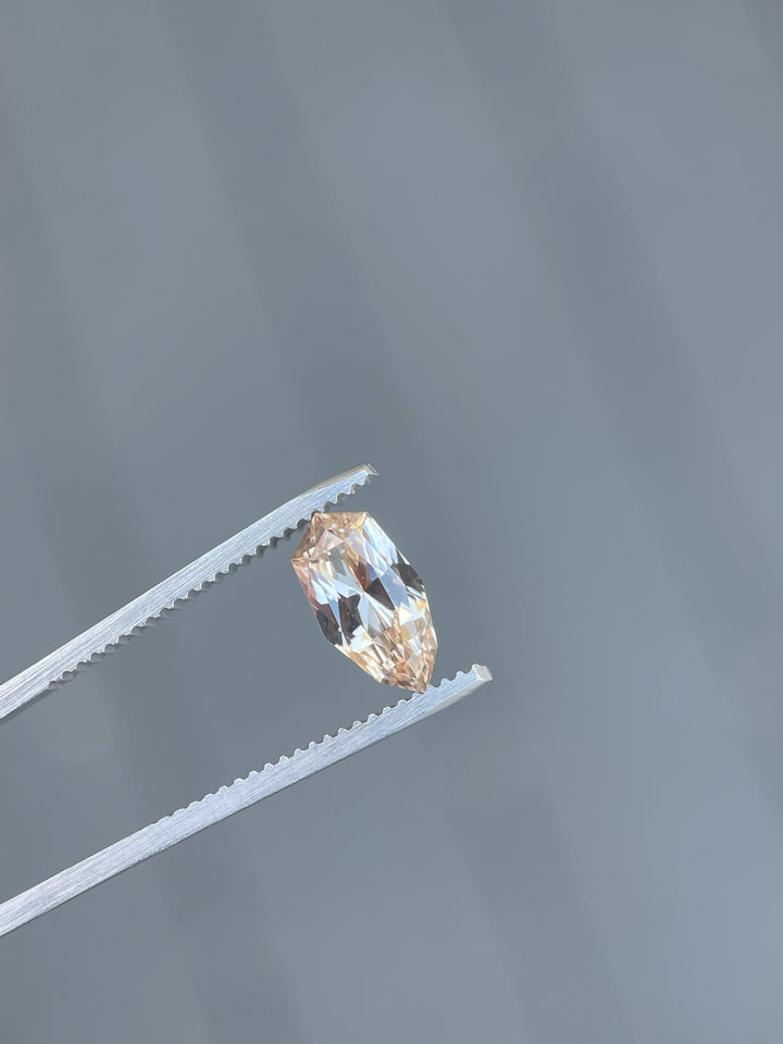 A 2.07-carat peach and champagne sapphire held by precision tweezers. The gemstone, cut in a custom geometric shield brilliant cut, shows its light peach and champagne hues under natural light. It measures 11.02mm by 5.87mm by 3.94mm and originates from Sri Lanka, untreated.