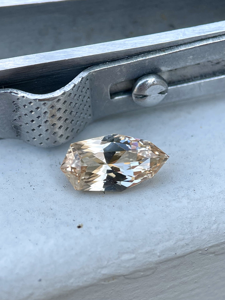 A 2.07-carat peach and champagne sapphire rests on a metal surface, showcasing its custom geometric shield brilliant cut. The gemstone’s sparkling facets catch the light, highlighting its unique shape and color. The stone is untreated and originates from Sri Lanka, with dimensions of 11.02mm by 5.87mm by 3.94mm.