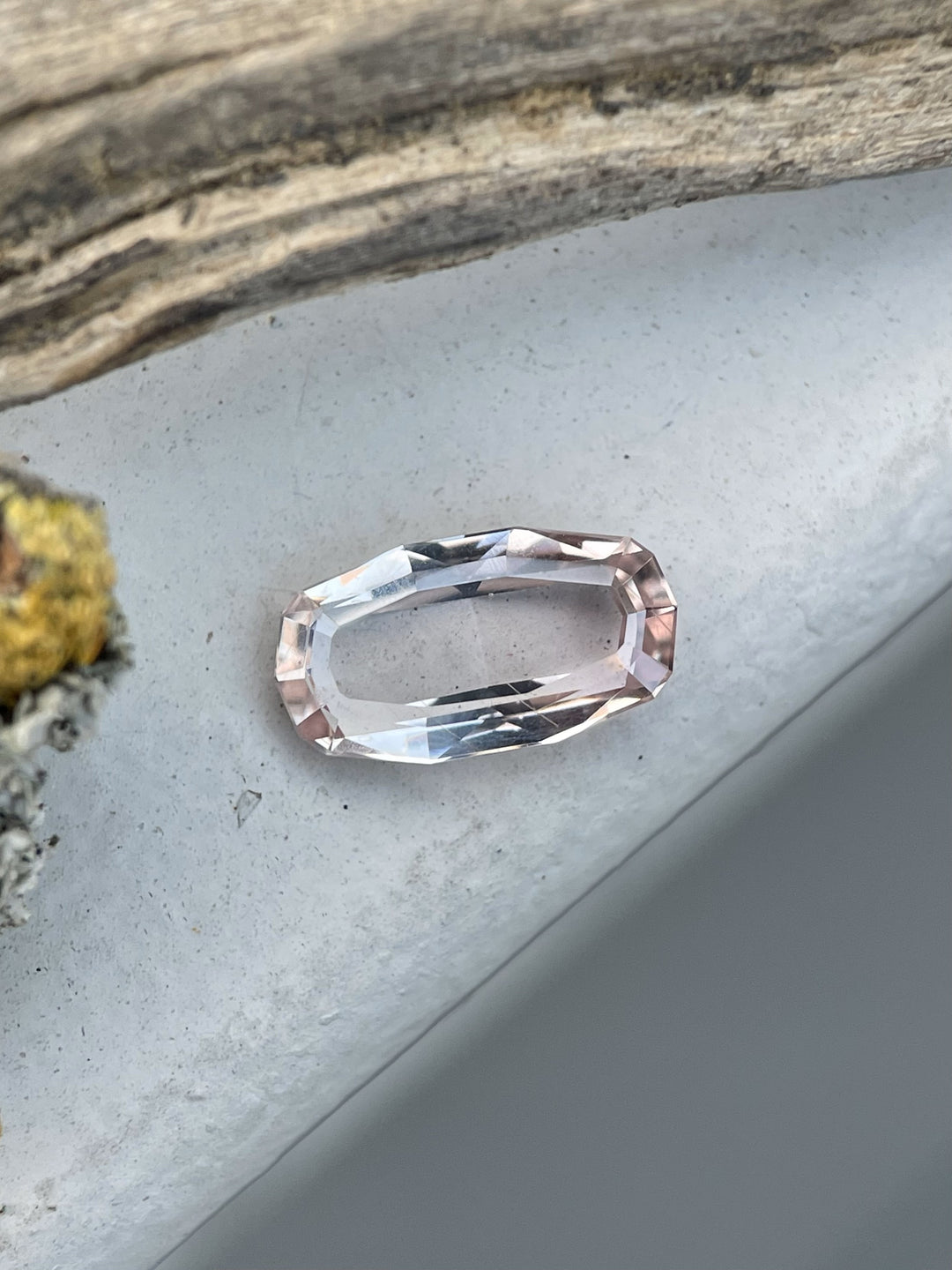 A 2.03-carat peach-colored sapphire in a custom elongated step-cut portrait style, resting on a rustic wooden surface. Dimensions: 11.08mm x 6.03mm x 2.74mm.