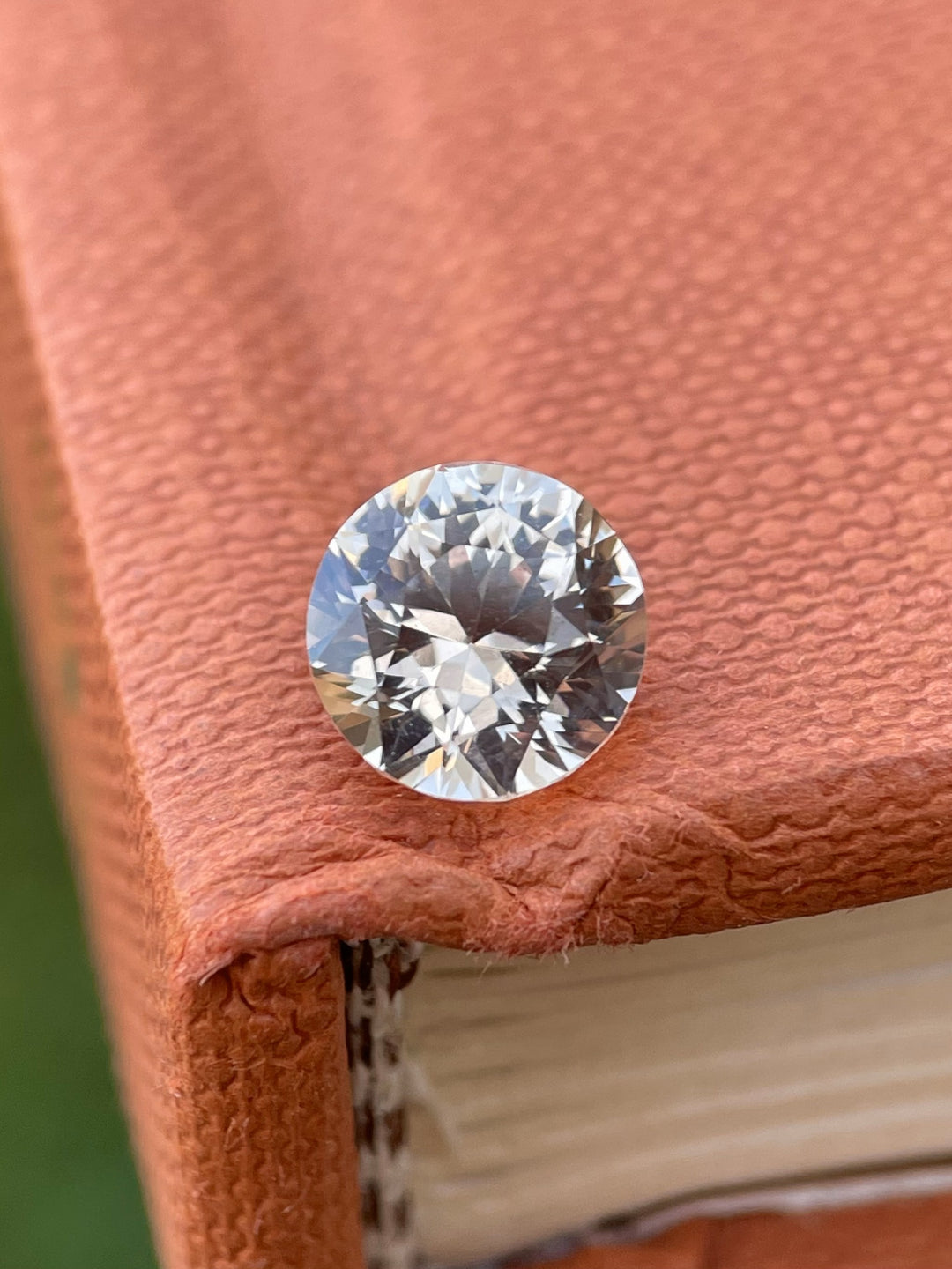 Close-up of a 2.57-carat peach sapphire resting on a textured book, showing off its fine round brilliant cut and soft, luminous peach hue.