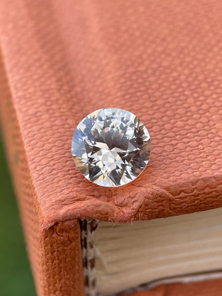 Close-up of a 2.57-carat peach sapphire resting on a textured book, showing off its fine round brilliant cut and soft, luminous peach hue.