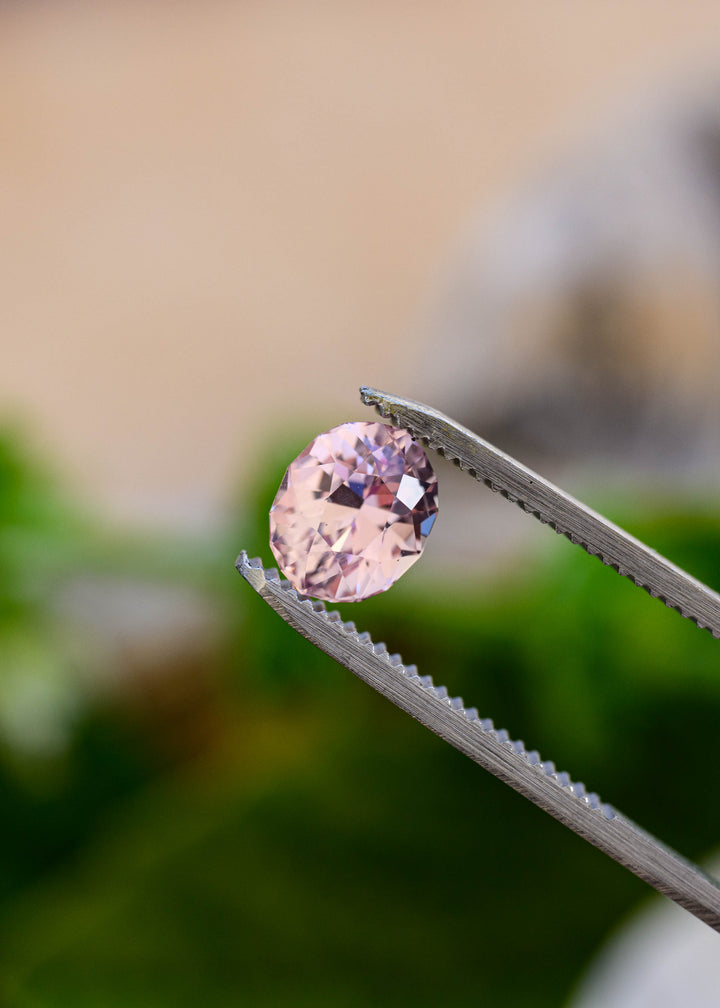 Peach blush pink Sri Lankan sapphire held in jeweler’s tweezers outdoors, displaying its custom geo oval brilliant cut and delicate pastel pink reflections.