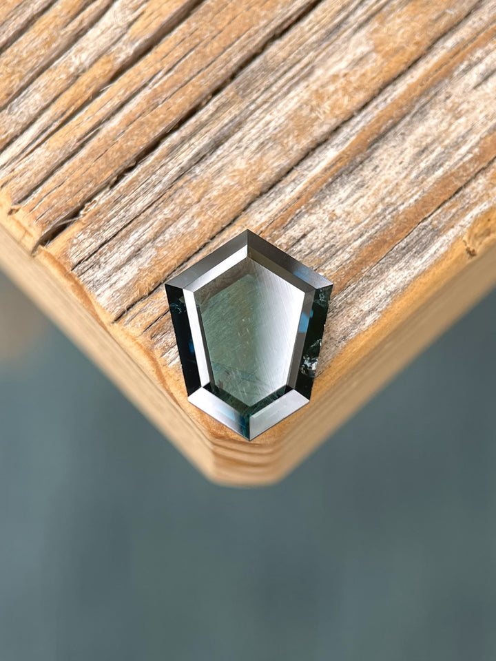 A 3.77-carat deep peacock teal sapphire with a custom shield portrait cut sits on a wooden surface. The gemstone displays a mix of blue and green hues, with sharp facets showcasing its clarity. Sourced from Garba Tula, Kenya, the stone is untreated and measures 12.54mm by 9.29mm by 3.02mm.