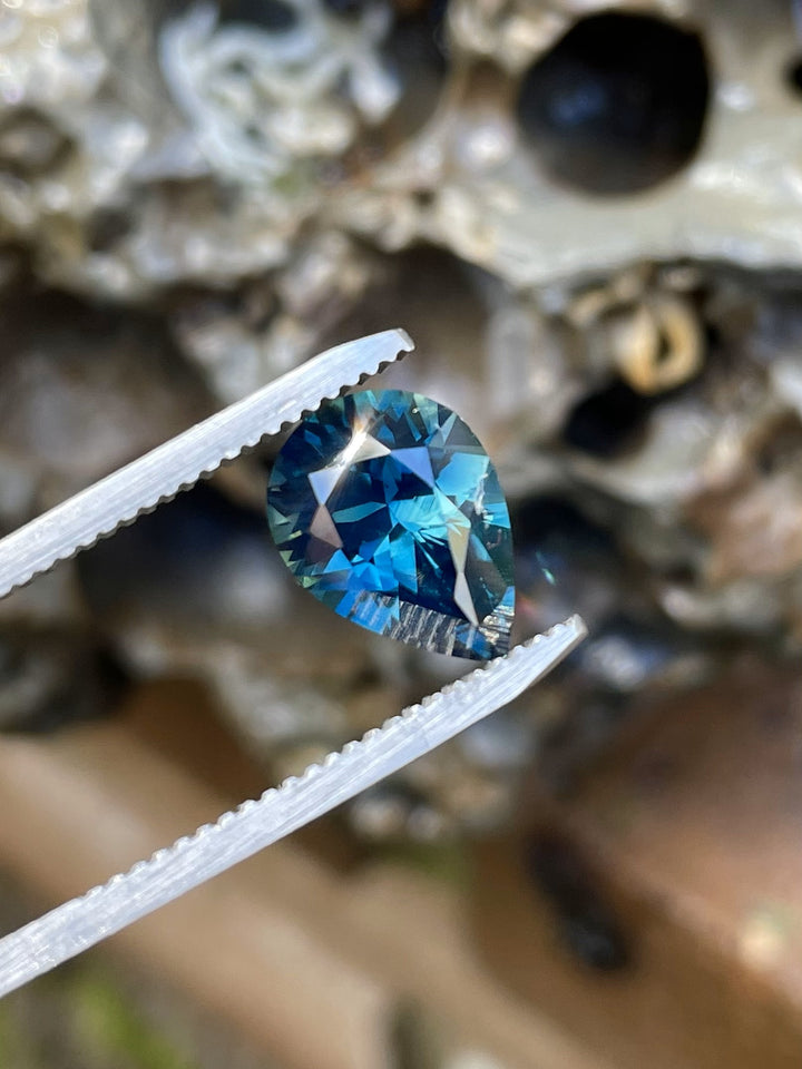 Close-up of a 4.20 carat pear cut sapphire held in tweezers, featuring vivid teal, blue, and green flashes against a textured backdrop.