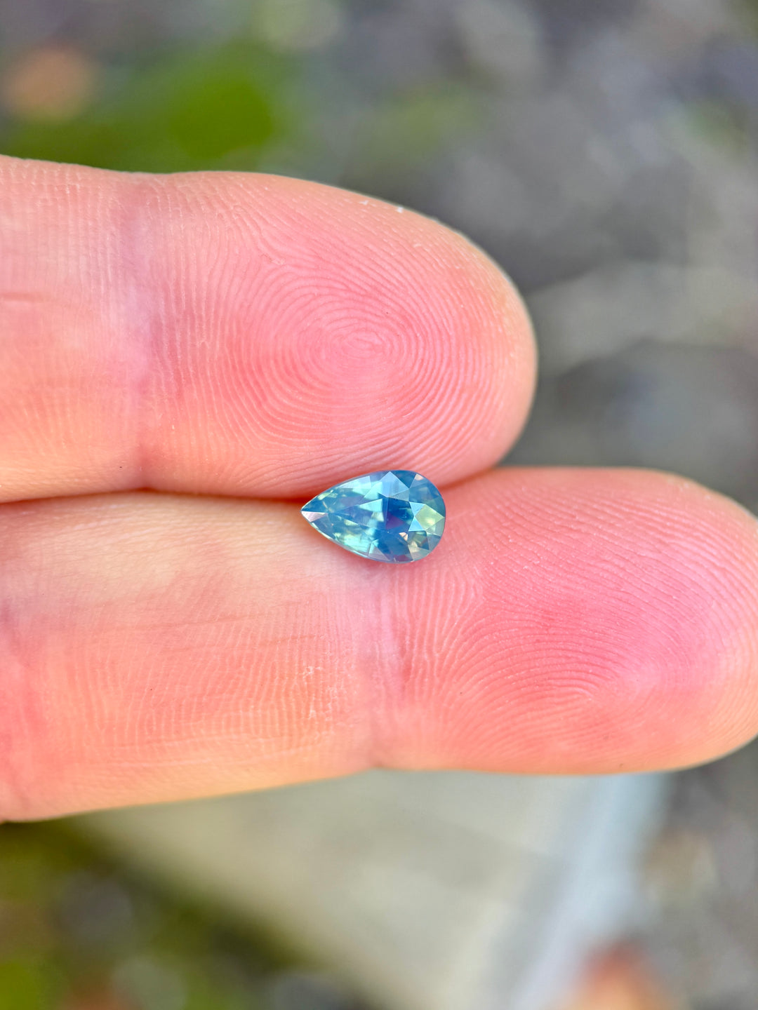Pear mixed cut sapphire resting on fingertips, showing layered teal, blue, and green tones with bright facet reflections.
