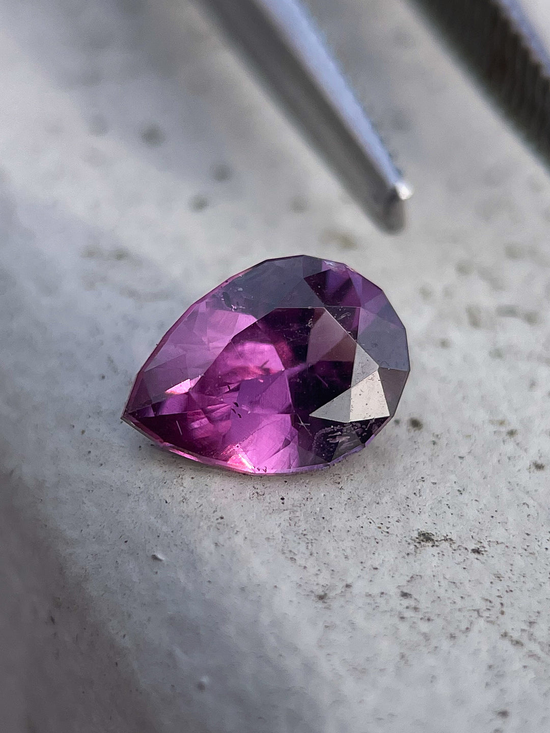 A modified brilliant pear-shaped sapphire with vivid pink and purple hues resting on a textured white stone surface.