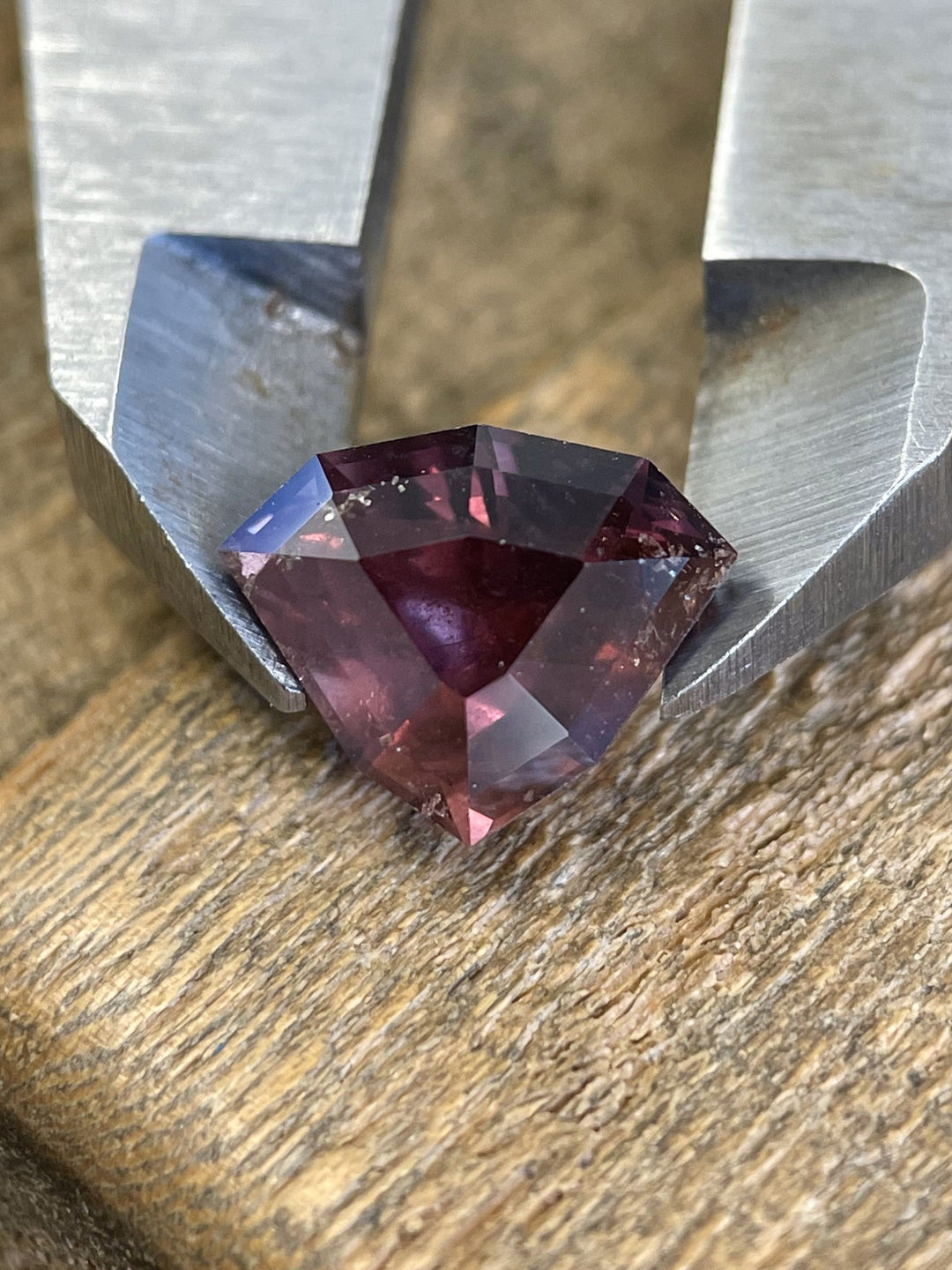 A 2.94-carat dusty rose mauve sapphire in a custom shield step cut, placed on a wooden surface with metal tweezers, showcasing sharp geometry and rich natural inclusions.