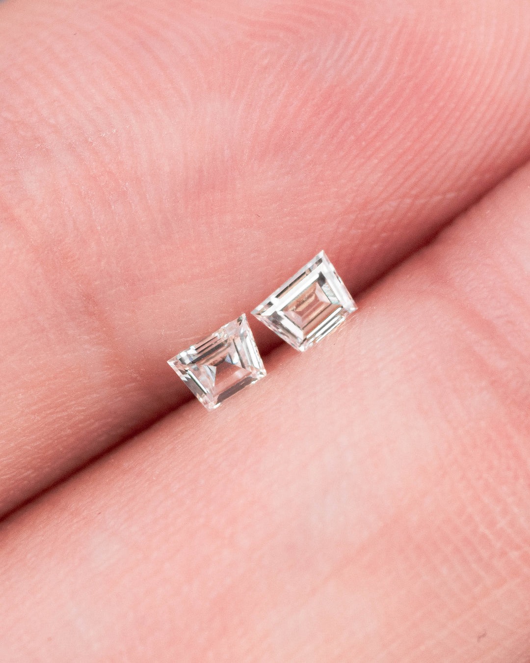 Matched pair of 0.18 carat F-G color VVS clarity Trapezoid step cut diamonds resting on fingertips, showing the size, brilliance, and clean faceting of the precision-cut South African diamond pair.