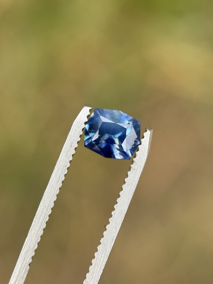 Unheated 1.60ct Madagascar parti sapphire held in gem tweezers outdoors, showing sky-denim and royal-blue flashes in a precision custom geometric cushion cut.