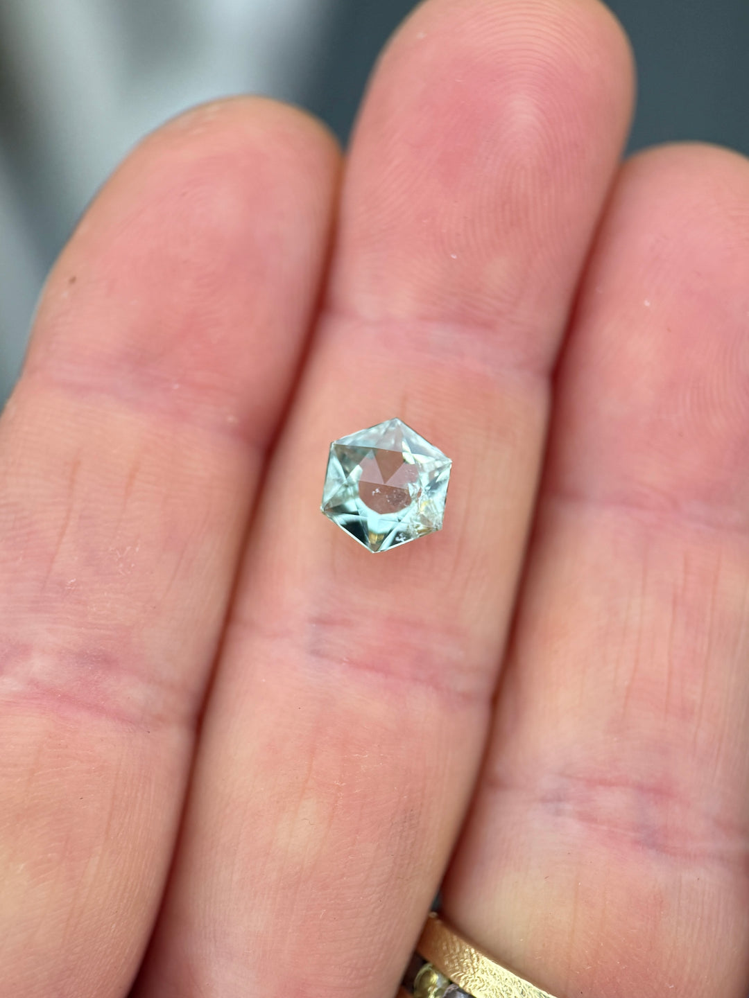 1.76 carat untreated Montana sapphire resting on fingertips, showcasing a precision custom hexagon mixed cut with mint sage and teal green color, large open culet, and rose cut table under natural light