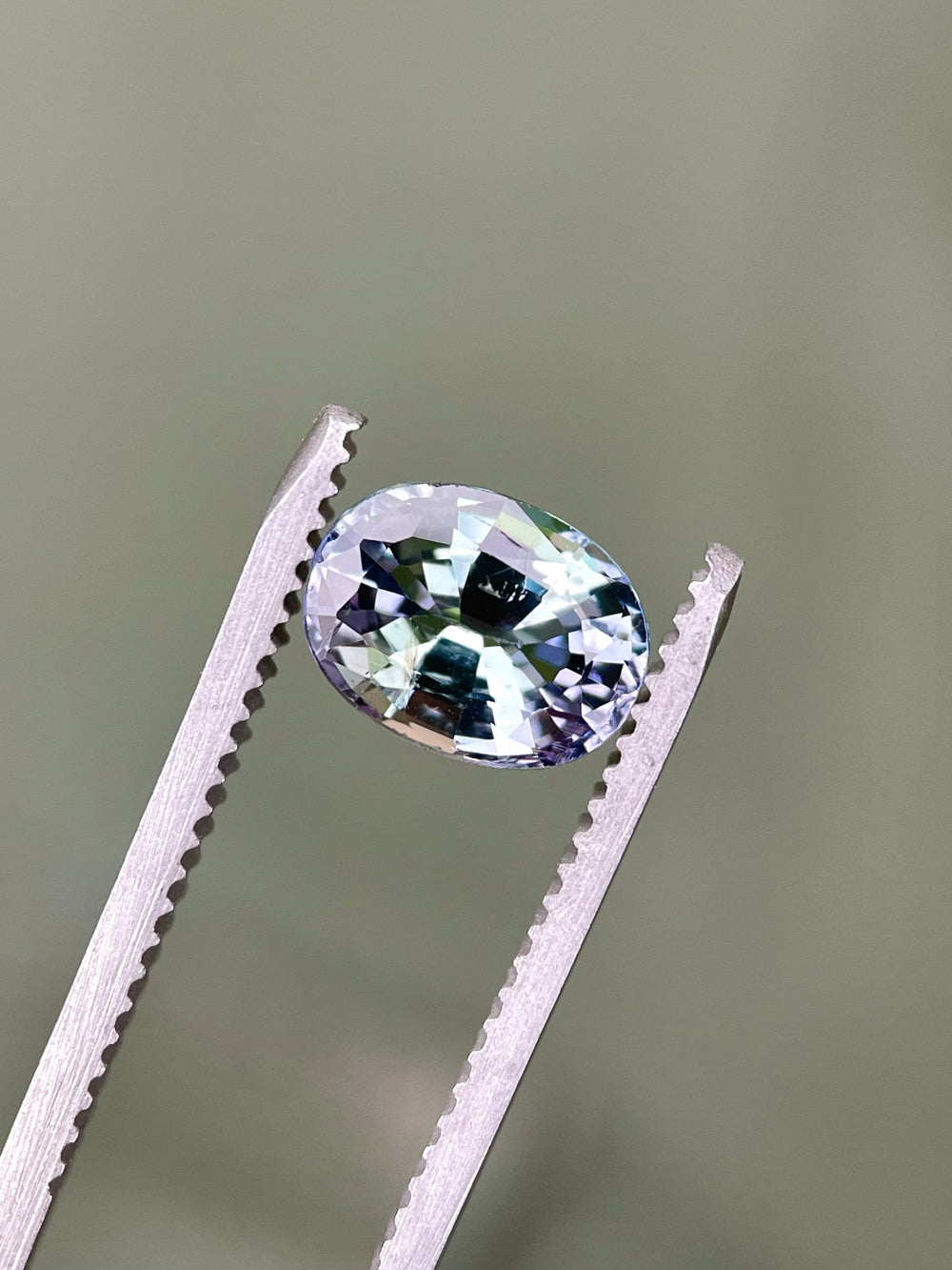 Close-up of a precision oval mix cut Tanzanite held in tweezers, highlighting its vivid green, teal, and purple flashes under soft lighting.