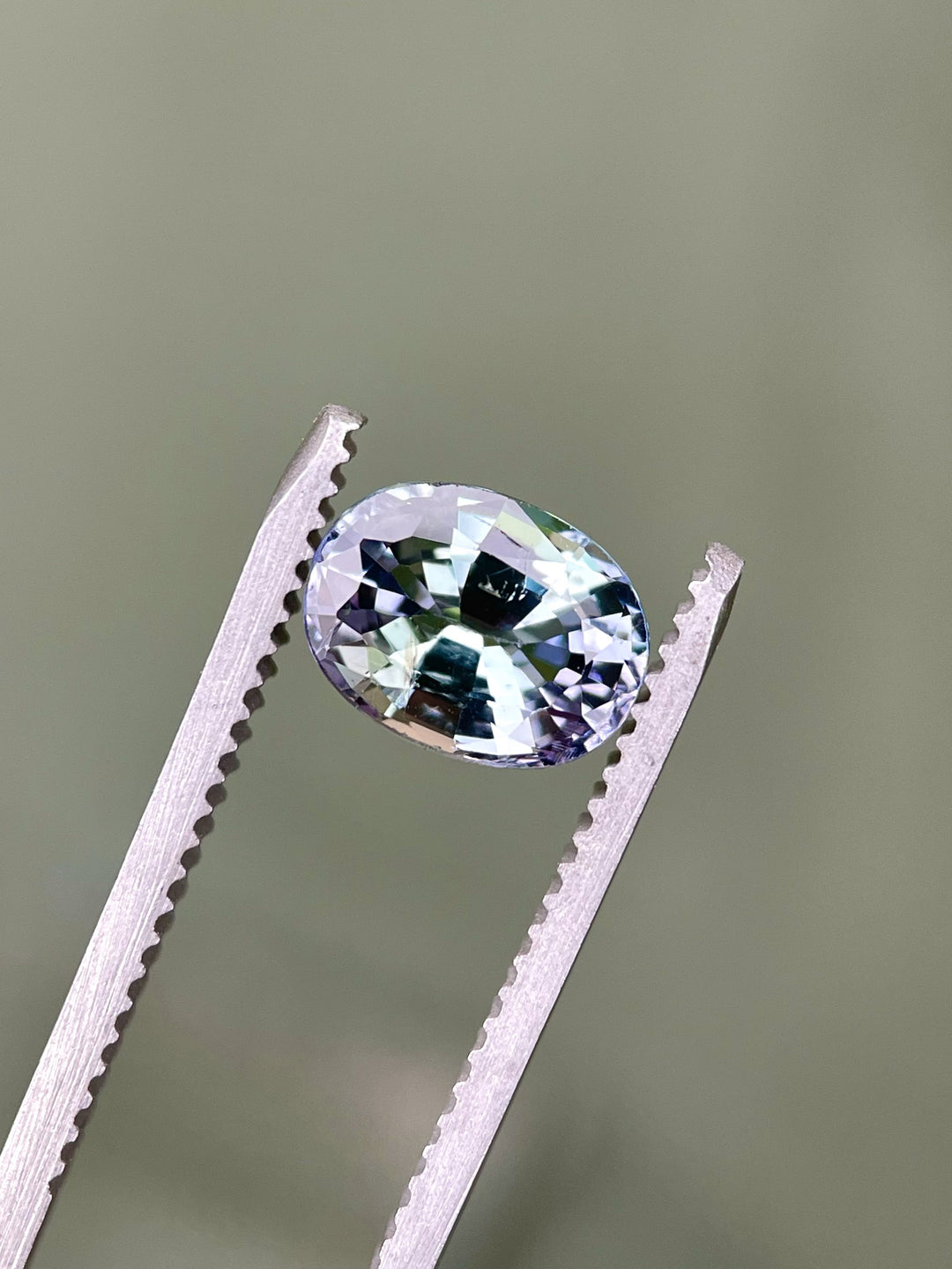 Close-up of a precision oval mix cut Tanzanite held in tweezers, highlighting its vivid green, teal, and purple flashes under soft lighting.