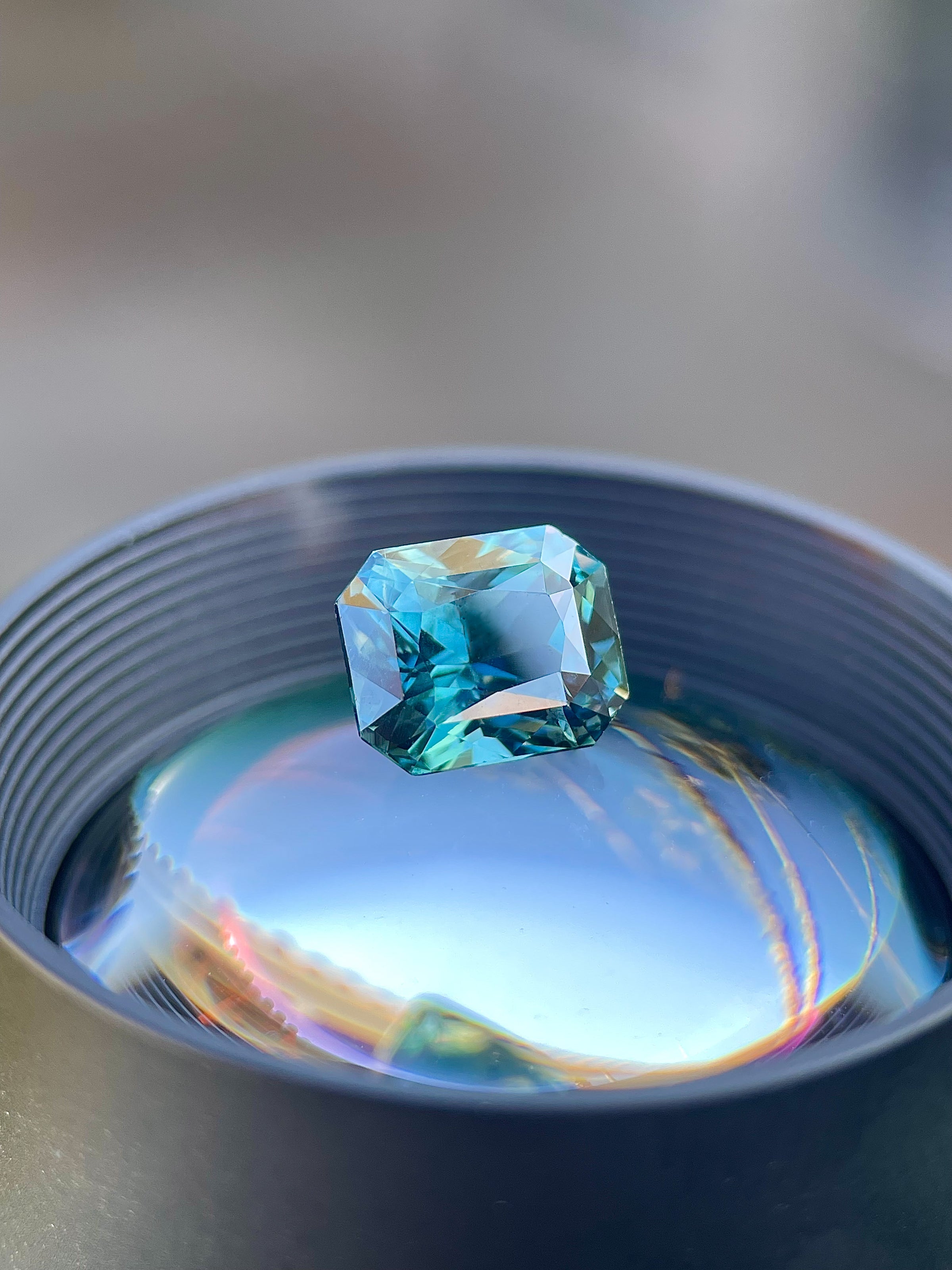 Precision cut teal sapphire on a gem lens, showcasing blue-green color, brilliance, and one-of-a-kind character for custom jewelry.