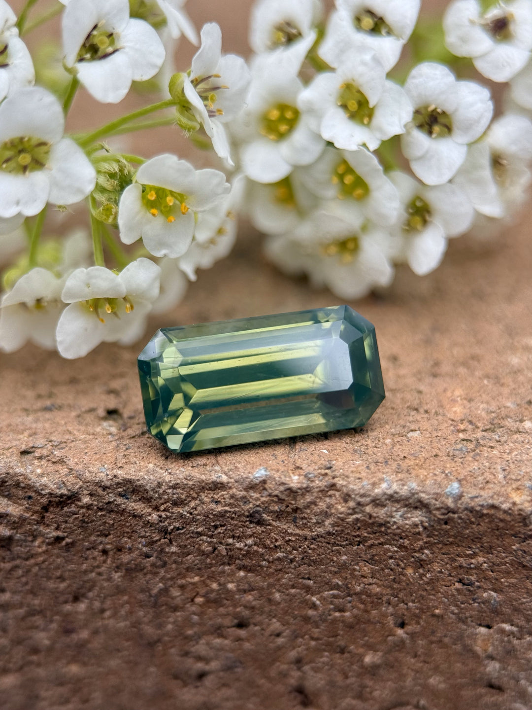 Close-up of a 3.36-carat precision-cut olive green sapphire resting near white flowers. The gem showcases an elongated emerald cut and glows with opalescent green hues.