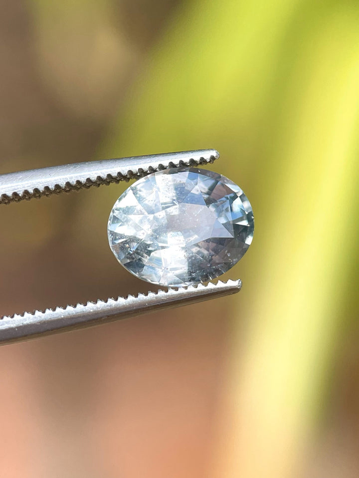 Close-up of a 3.19 carat sapphire with a mix brilliant oval cut, held in tweezers, showcasing its unique teal and seafoam color change properties, perfectly cut to capture light.