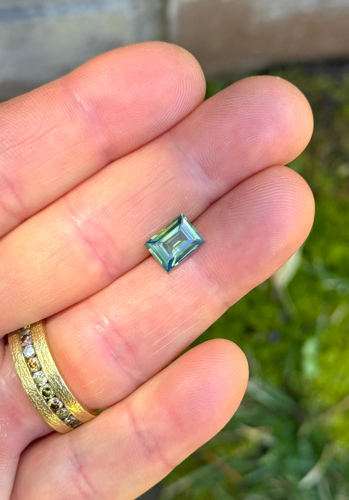 Precision baguette step cut sapphire resting on fingertips, showing layered teal, blue, and green tones with reflective linear facets.