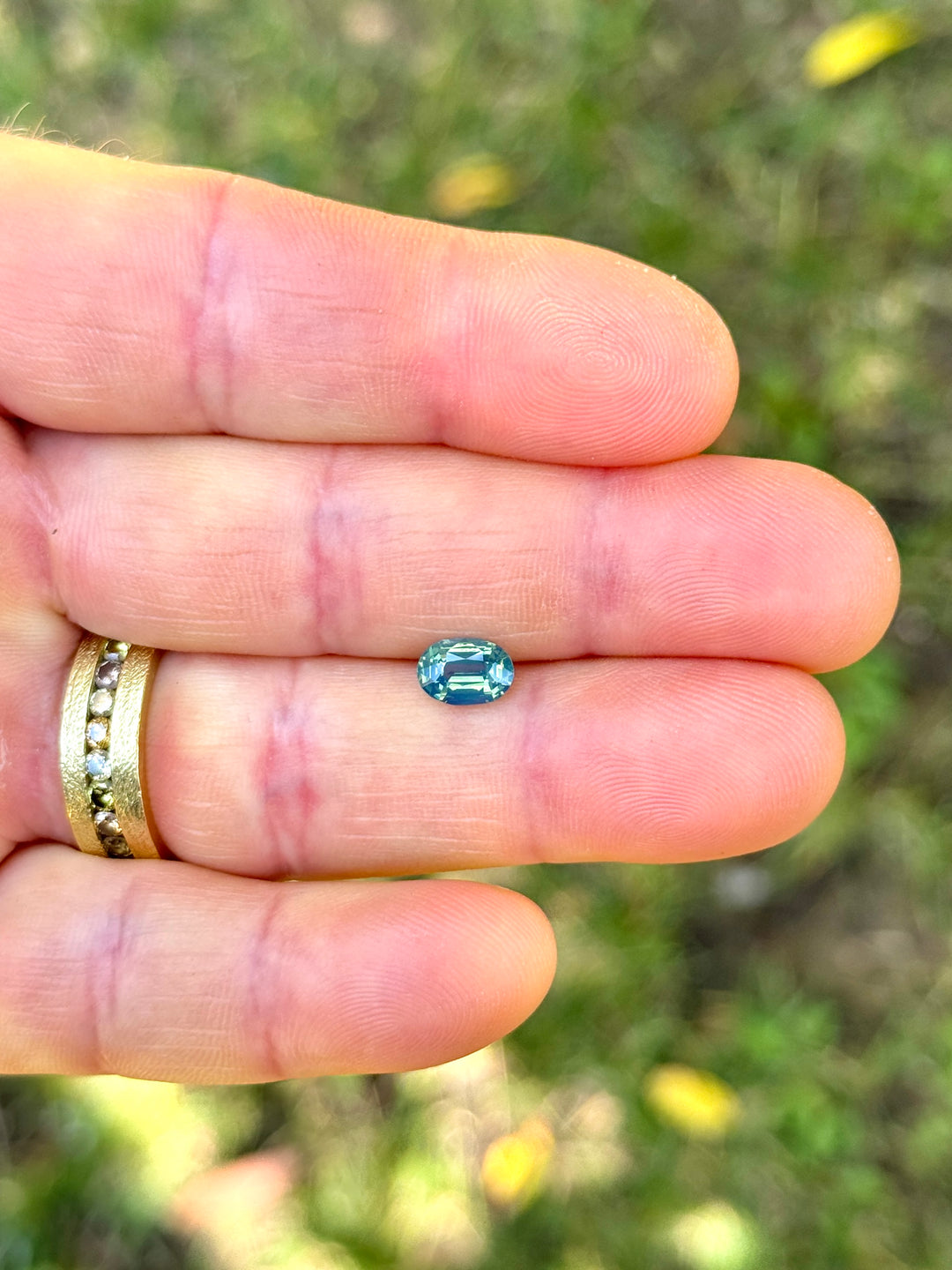 Precision elongated oval sapphire resting on fingertips, showing layered teal, blue, and green tones with bright facet reflections.