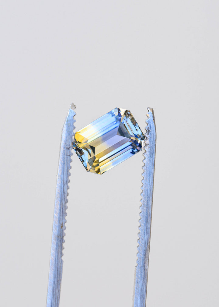 Precision emerald-cut Sri Lankan parti sapphire held in jeweler’s tweezers, displaying crisp clipped corners and distinct bands of periwinkle blue and pale golden yellow.