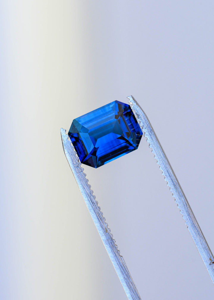 Precision emerald-cut royal blue sapphire held in jeweler’s tweezers against a pale background, displaying its rectangular shape, clipped corners and intense blue saturation.