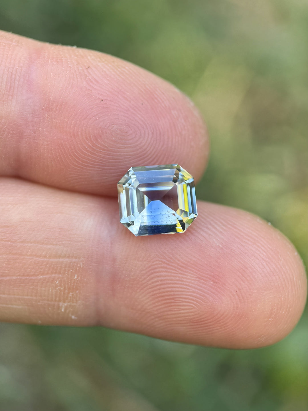 Light yellow and blue parti sapphire weighing 4.04 carats shown on fingertip for size reference, highlighting the precision emerald cut step facets and Madagascar origin.