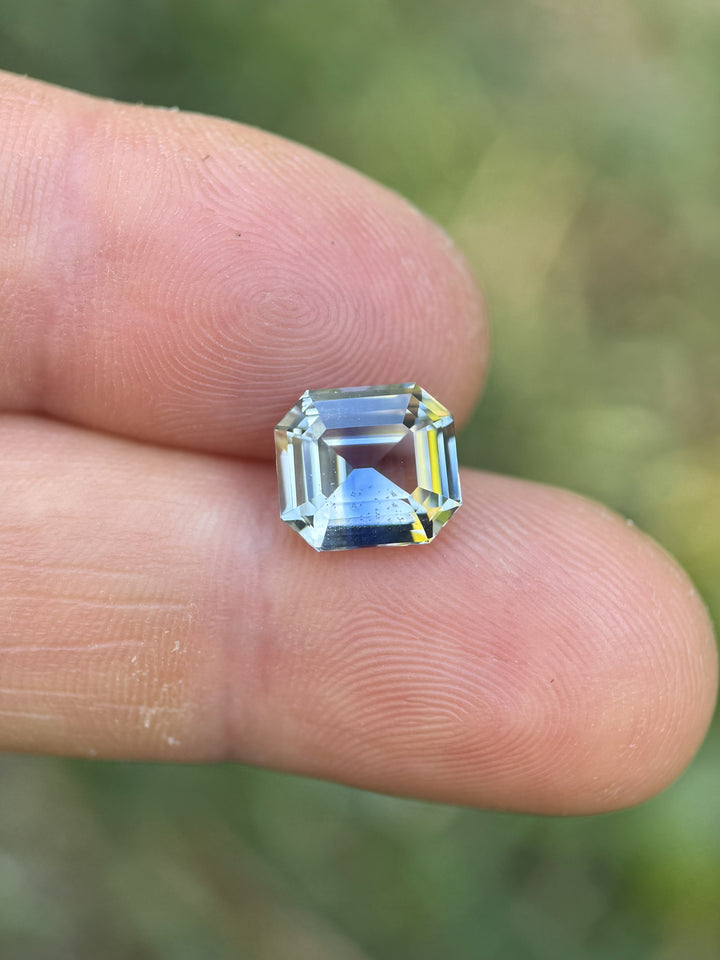 Light yellow and blue parti sapphire weighing 4.04 carats shown on fingertip for size reference, highlighting the precision emerald cut step facets and Madagascar origin.