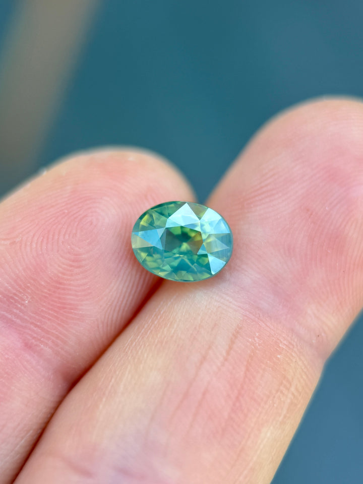 Precision oval mixed cut sapphire resting on fingertips, showing opalescent teal, blue, and green flashes with excellent depth and saturation.