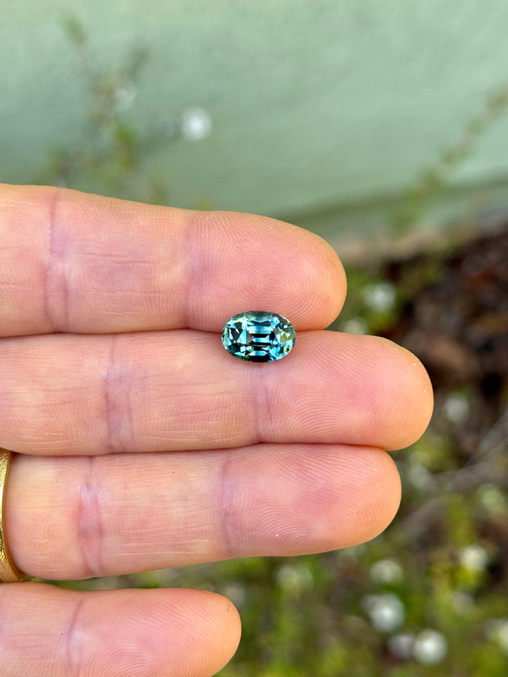 Precision oval mixed cut sapphire resting on fingertips, showing vivid teal, blue, and green flashes across the crown facets.