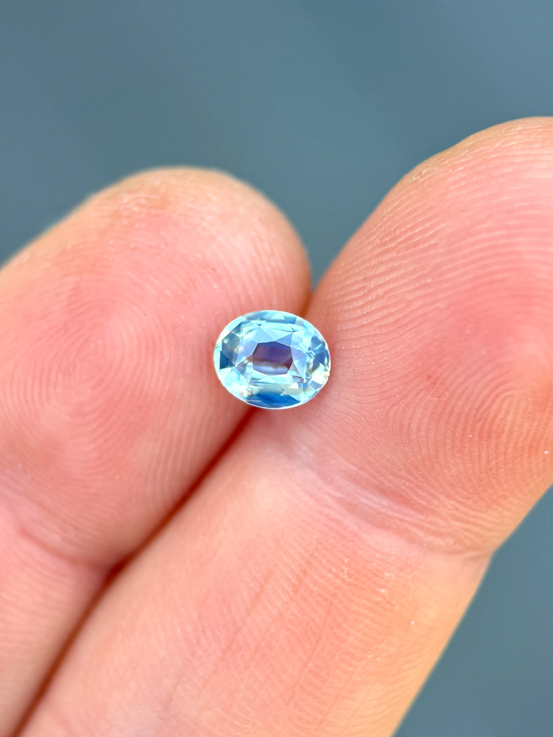 Precision oval cut sapphire resting on fingertips, showing powder blue color with delicate silvery reflections and bright brilliance.