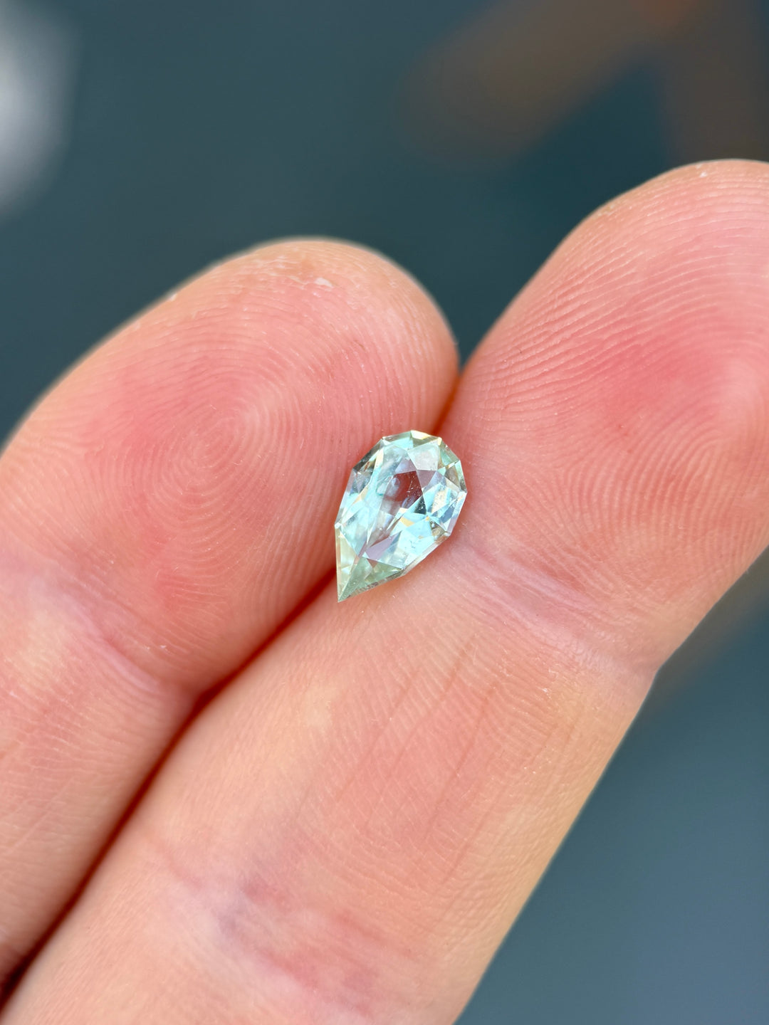 Precision pear cut sapphire resting on fingertips, showing light seafoam green color and lively facet reflections.