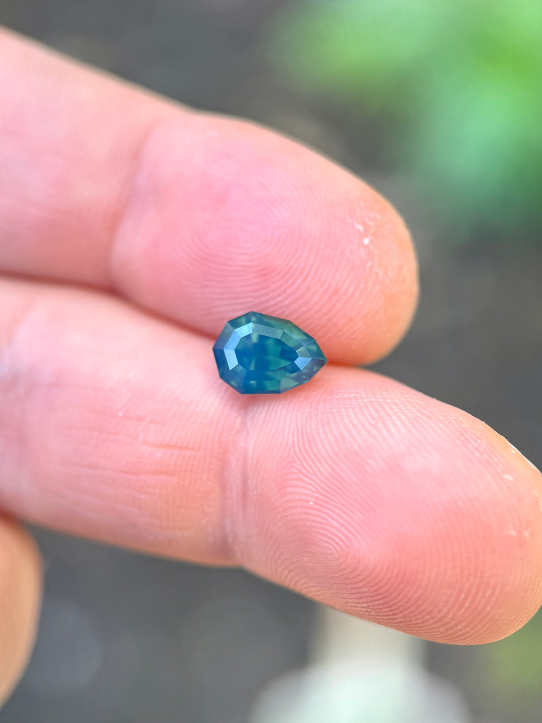 Precision shield step cut sapphire resting on fingertips, showing layered teal, blue, and green tones with broad mirror-like reflections.