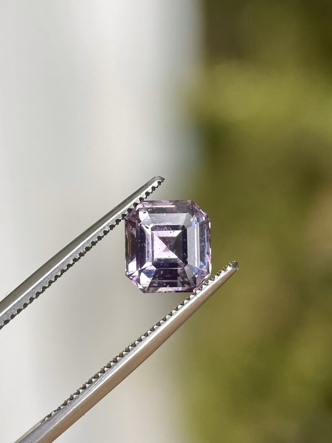 A 2.60-carat purple and pink sapphire with a modified Asscher cut is held by tweezers. The sapphire's precision cut and natural hues are showcased against a blurred natural background. The gemstone, untreated and no heat, measures 7.71mm by 7.25mm by 4.97mm.