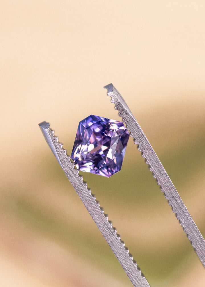 Loose precision radiant cut sapphire held in tweezers, displaying violet and pink tones and crisp facet reflections from Madagascar.
