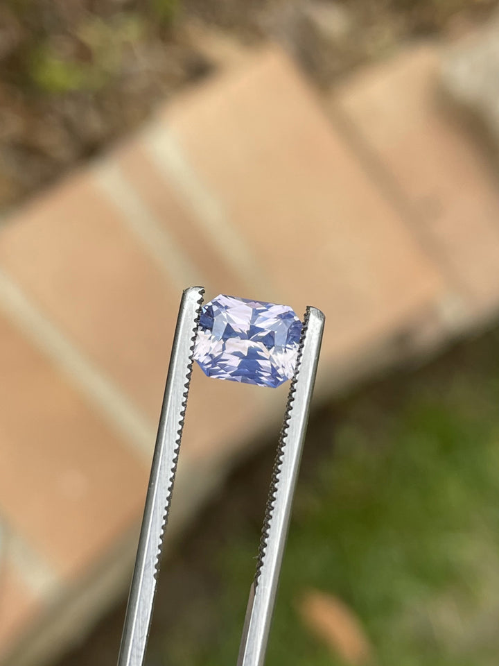 Close-up of a radiant cut 2.16-carat Sri Lankan sapphire in natural light, showing off its pastel peach and lavender sparkle between gem tweezers.