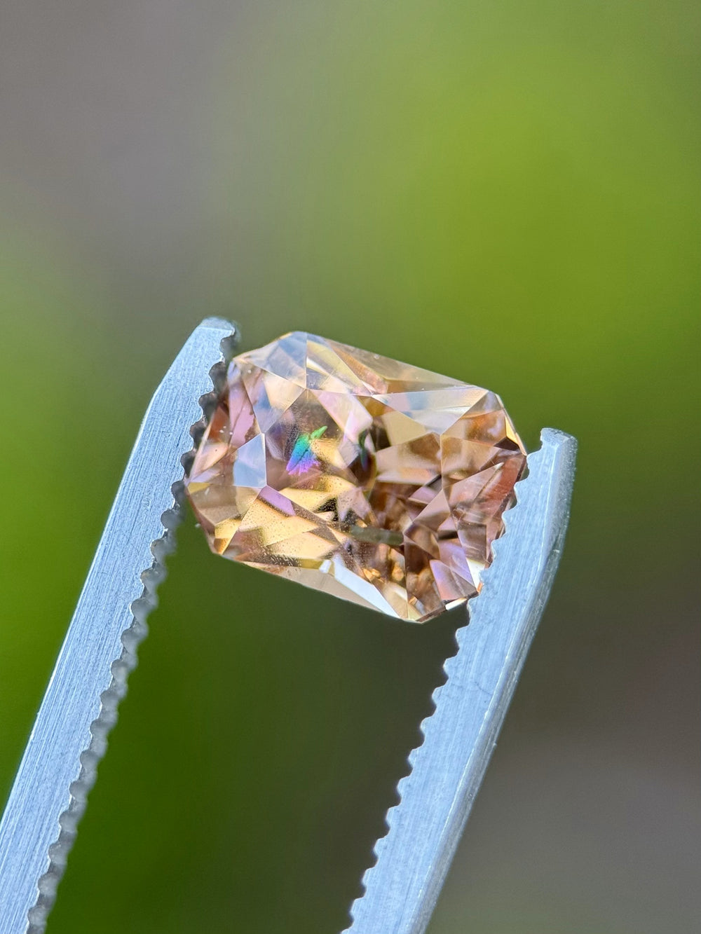 Custom modified radiant Tanzanite held in tweezers with a soft green background, highlighting its untreated purple, peach, and champagne color blend.