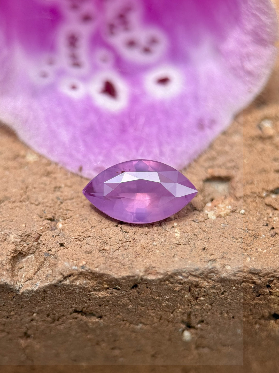 1.15CT marquise sapphire in pink-violet hues placed on stone with purple flower in background, highlighting its untreated natural beauty