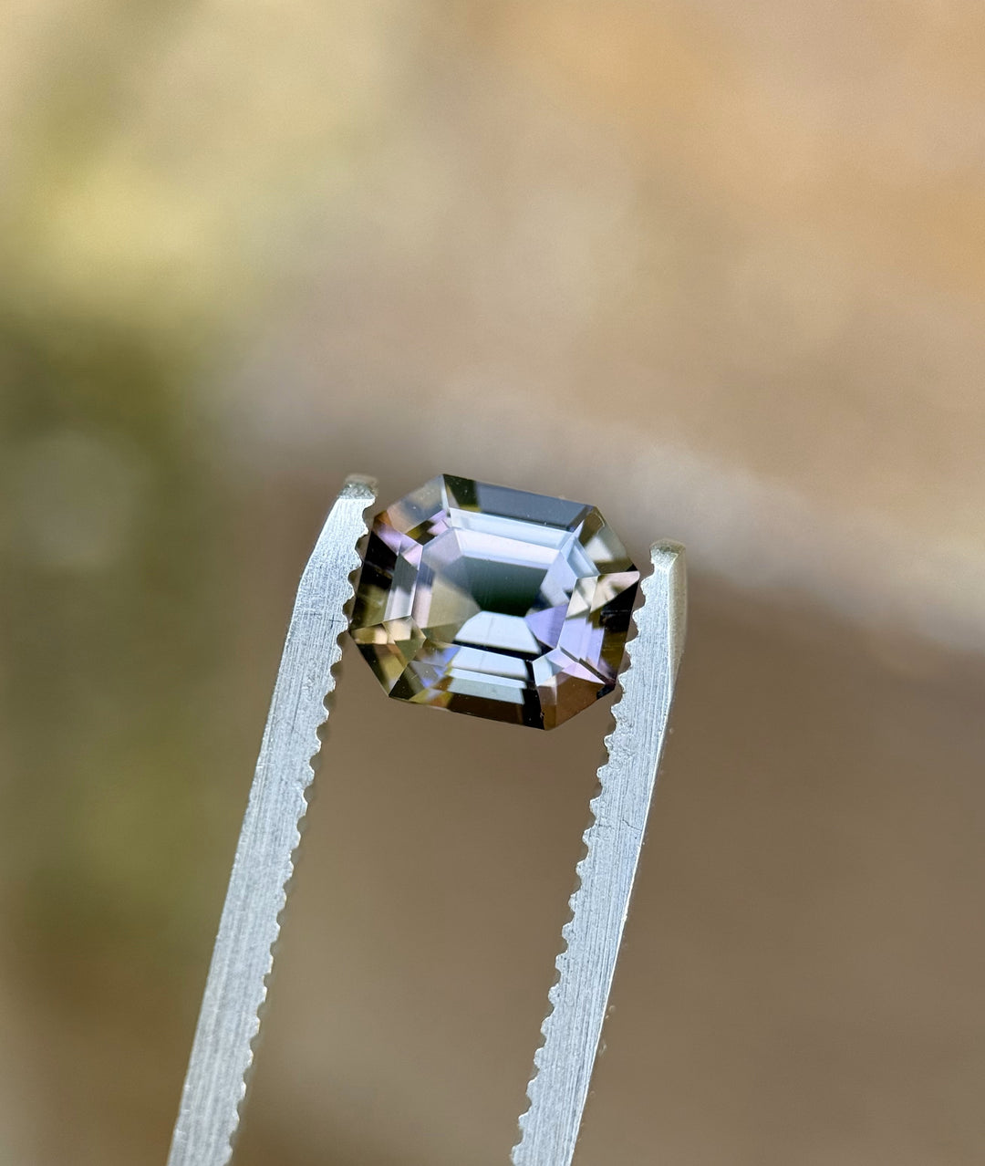 1.32 carat emerald cut Tanzanite gemstone in mermaid, teal, and purple tones, displayed in precision tweezers against a soft background.