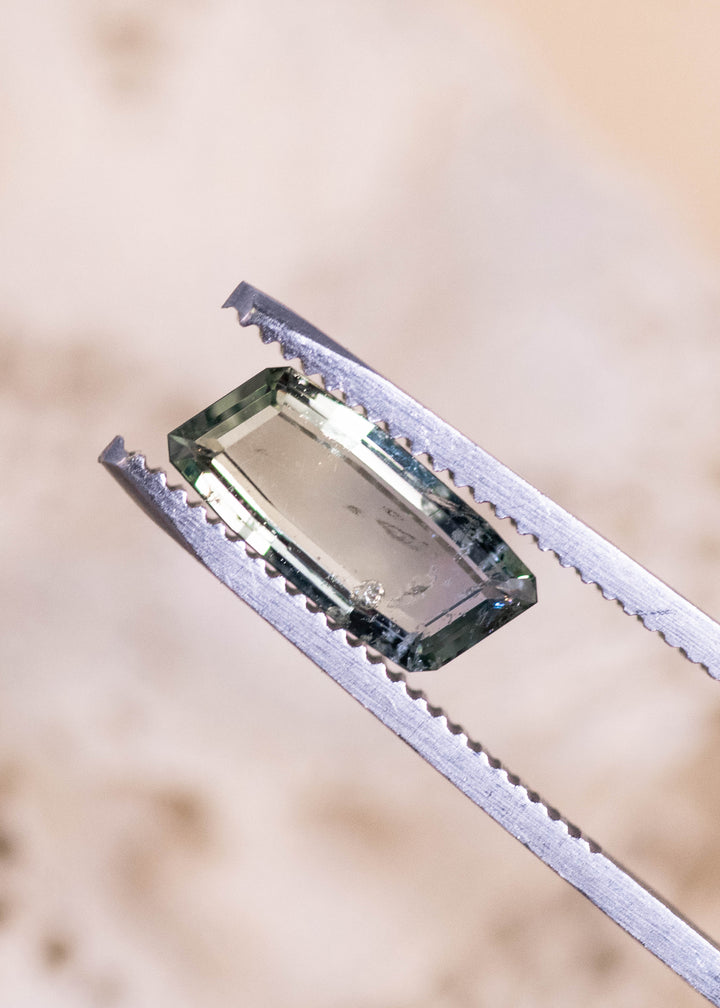 2.31 carat yellow green teal sapphire held in gem tweezers, displaying precision step-cut facets and elongated baguette portrait shape from the Umba River, Tanzania, untreated