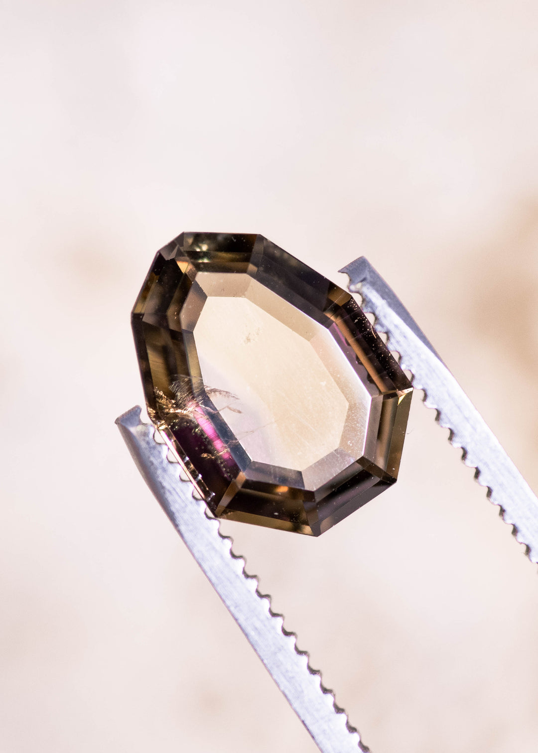 Custom shield portrait cut Tanzanite held in gem tweezers, showing crisp geometric facet structure and pink-purple-peach tones in a 3.20 carat untreated Tanzanian gemstone