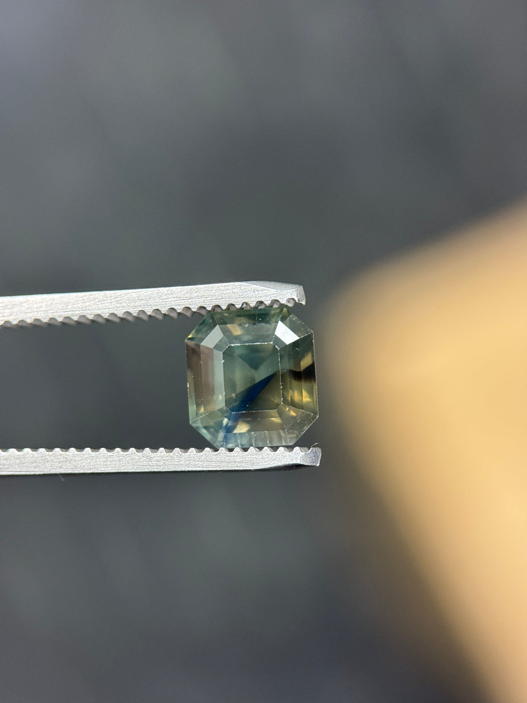 Precision emerald cut 3.40CT Nigerian sapphire held in tweezers, showcasing opalescent olive teal green color zoning and natural sapphire clarity